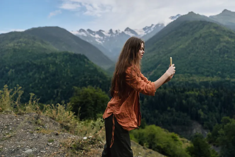 Woman standing on a hillside taking a selfie with a smartphone amid majestic mountains and lush landscape showcasing outdoor exploration