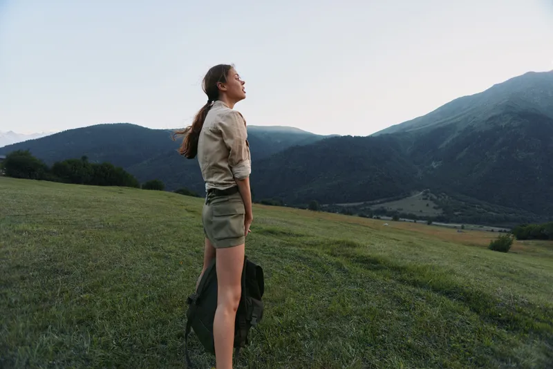 Young woman standing on a green hill looking at mountains during sunset. Outdoor lifestyle concept with nature, freedom, and adventure in soft natural light.