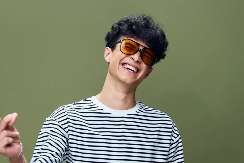 Young man laughing joyfully wearing sunglasses and striped shirt against soft green background, capturing happiness and casual style in modern portrait photography.