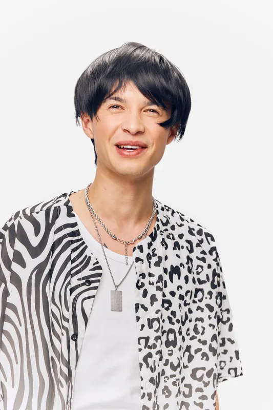 young man wearing patterned black and white shirt with layered silver necklaces smiling happily against a plain white background, casual style and joyful expression