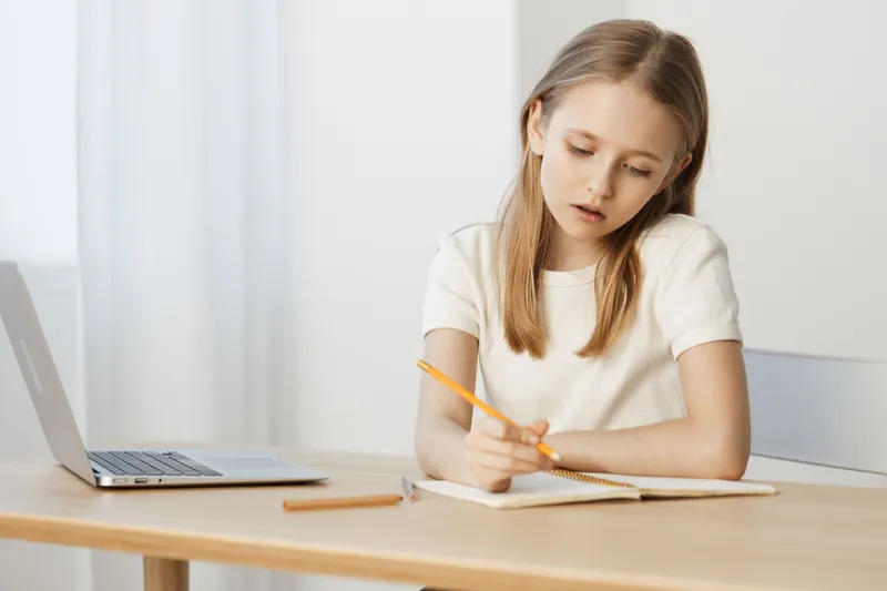 Young girl studying at a desk with a laptop, focusing on her notes, wearing a casual white shirt, showcasing concentration and determination in a bright, minimalistic setting