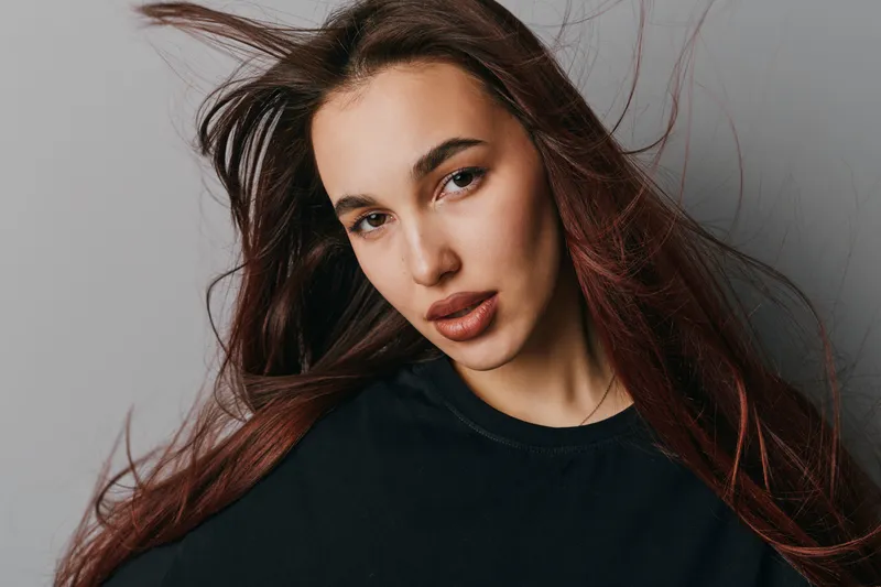 Woman with long hair and natural makeup wearing black top posing against gray background. Portrait of confident female with loose hair and serious expression in studio lighting.