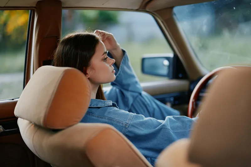 Young woman with long brown hair sitting in a vintage car, wearing a denim jacket, looking pensive against a blurred green background.