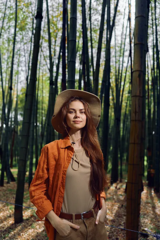 Woman bamboo forest hat portrait nature outdoors longhair, wearing straw hat and orange jacket, hands in pockets with confident smile, sunlit trees and peaceful foliage for travel and lifestyle.