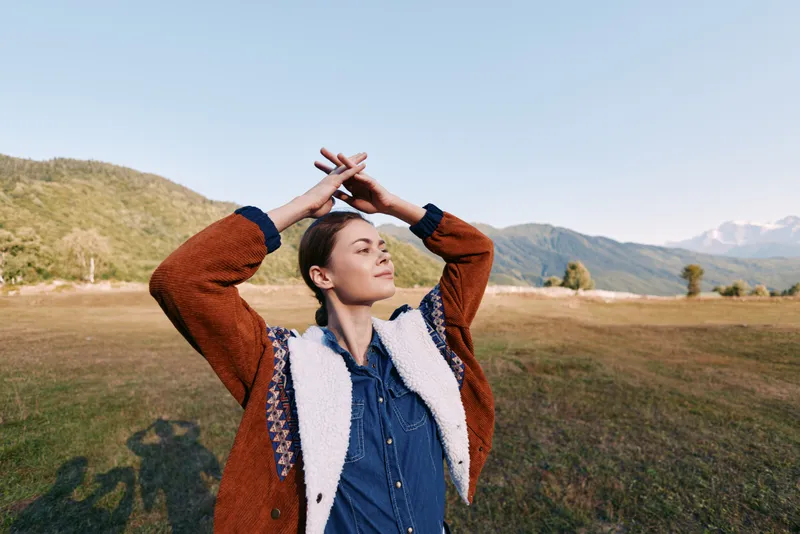 Woman portrait in nature among mountains and open field, relaxed and eyes closed outdoors wearing a cozy sweater, enjoying sunlight, breeze and peaceful rural scenery on a sunny day.