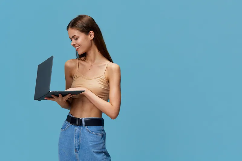 Smiling woman holding laptop with casual beige top and blue jeans, standing against plain blue background, showcasing technology and relaxed mood.