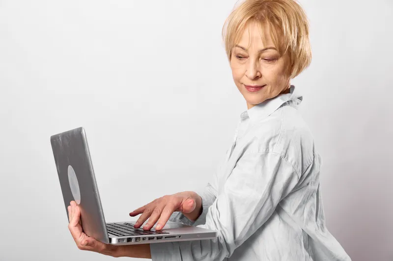 Elderly woman holding laptop indoors, typing on keyboard, calm smile, focused portrait, home setting, modern technology, relaxed posture, soft neutral light