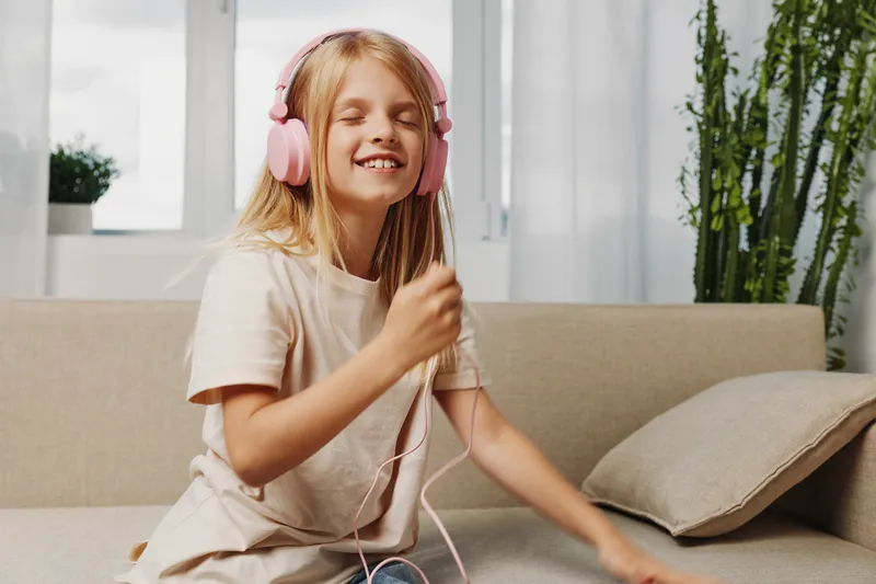 Smiling girl with long blonde hair wearing pink headphones enjoys music with closed eyes, sitting on beige sofa in bright living room with natural light and plants.