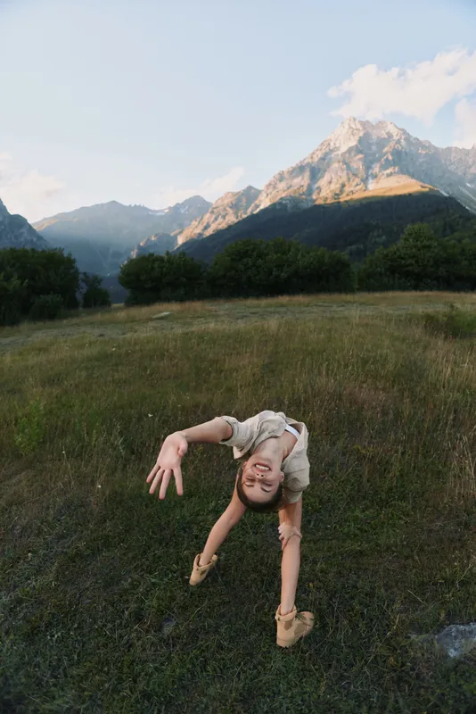 Young woman performing a backbend in an open meadow with distant mountains, soft daylight, and lush grass, conveying fitness, outdoor activity, balance and freedom