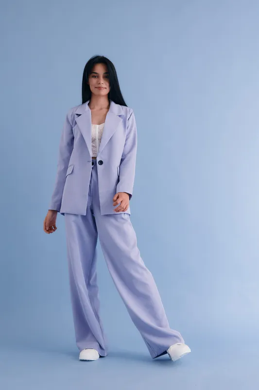 young woman in lilac pantsuit posing confidently on blue background, wearing casual white shoes and delicate lace top, standing full-length with relaxed expression