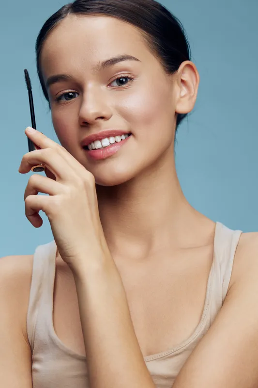 Young woman with a natural look holding an eyebrow pencil, smiling on a soft blue background, showcasing beauty, confidence, and wellness in a minimalist style