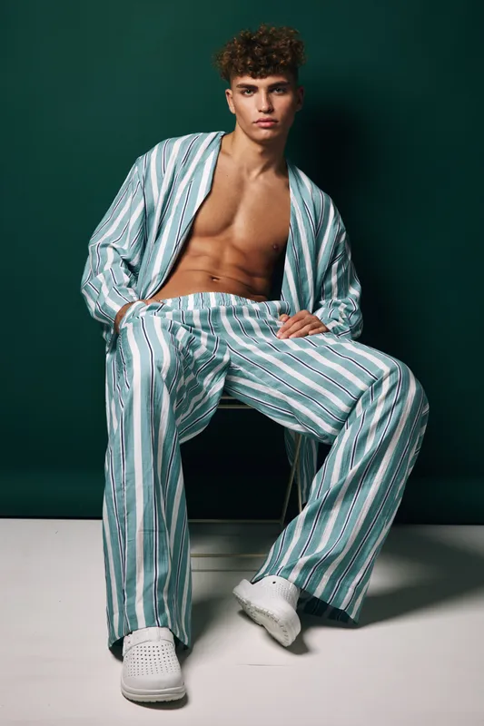 Young man wearing striped pajamas sitting in chair with hands in pockets and casual pose against green background. Relaxed shirt and shoes added leisure vibe to scene.