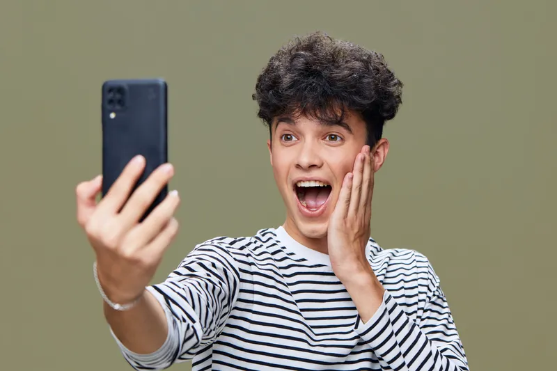 Excited young man taking a selfie with smartphone, wearing striped shirt and showing surprised facial expression against green background in casual indoor setting.
