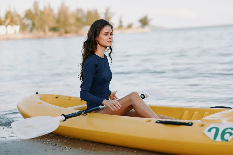 Young woman sits gracefully in a yellow kayak by the water, wearing a navy long sleeve top, relaxed pose, calm shoreline, soft light, gentle waves, outdoor adventure with reflective mood.