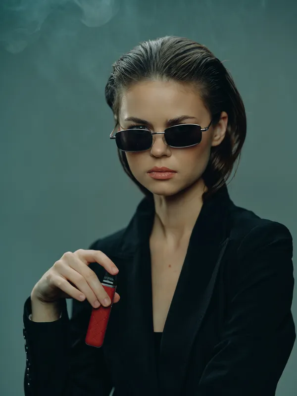 Stylish woman in black sunglasses and jacket holding a red vape device with smoke on dark background