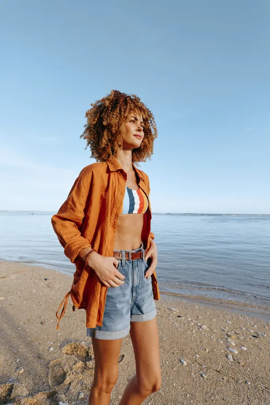 Woman stands on sandy beach by calm ocean horizon wearing rust jacket and denim shorts curly hair summer fashion lifestyle relaxation soft daylight scenic seascape outdoor travel vacation