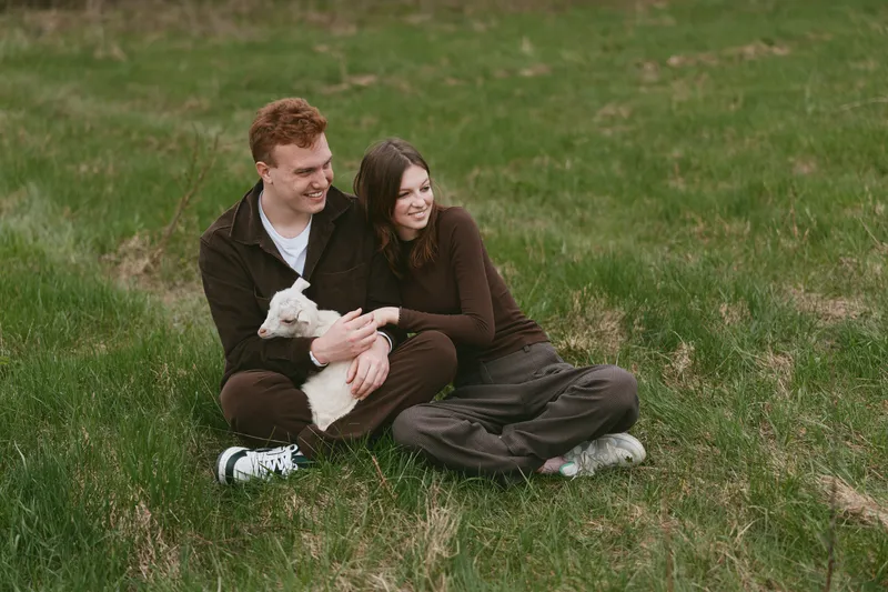 Couple outdoors with a goat kid in a field, showing affection and relaxed lifestyle, capturing nature, grass, and smiling joy in a warm, candid moment.