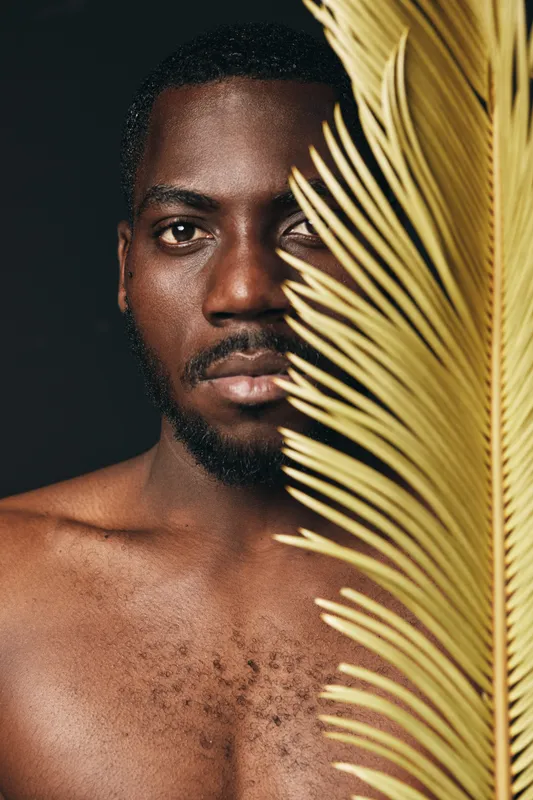 African man with dark skin and beard in creative conceptual visual style, half face covered by yellow palm leaf, closeup portrait with artistic lighting and natural texture on black background.