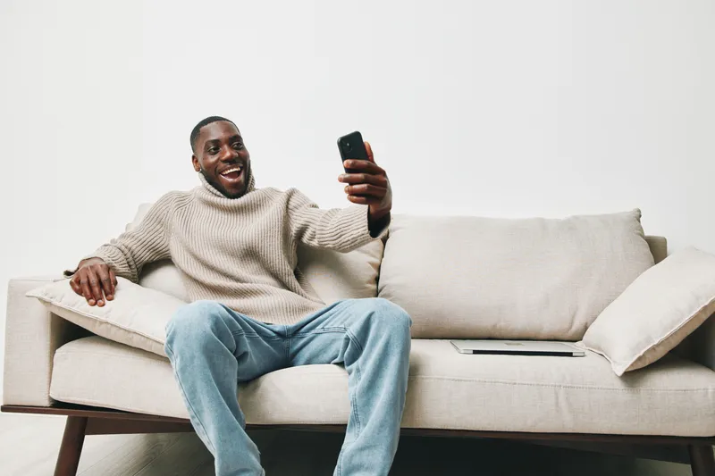 A joyful African man in a cozy sweater taking a selfie on a couch, showcasing happiness, comfort, and modern lifestyle in a minimalistic setting