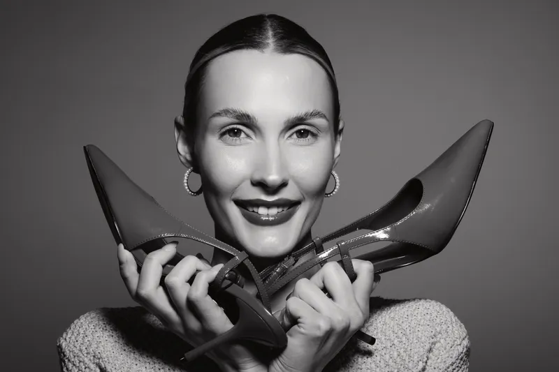 Happy caucasian woman with light skin and dark hair holds glossy high heel shoes close to face. Studio portrait with focus on fashion and style concept.