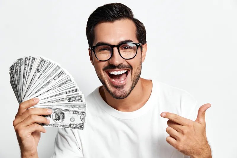 Smiling young man holding stack of money, showcasing wealth and happiness, dressed casually with glasses, isolated on plain white background.