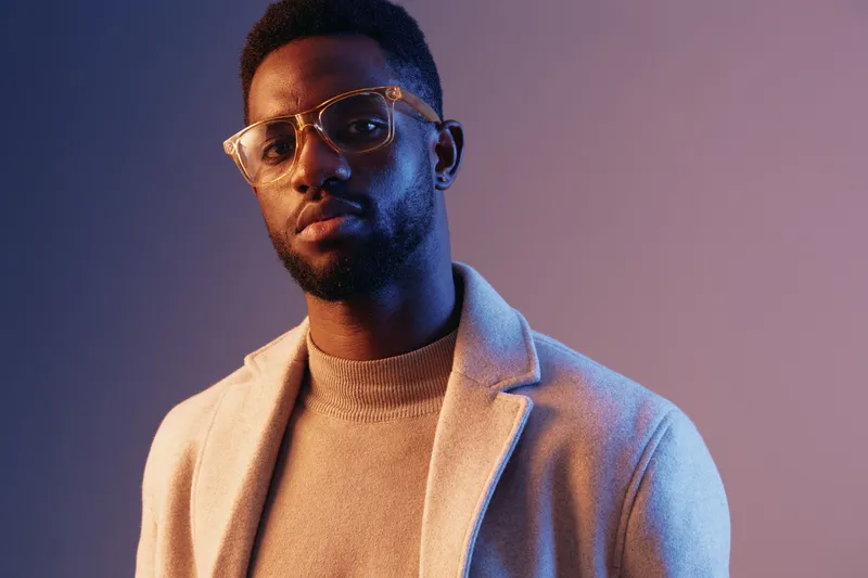 Stylish young Black man wearing glasses and a beige coat, exuding confidence and sophistication against a gradient background, perfect for fashion or lifestyle concepts