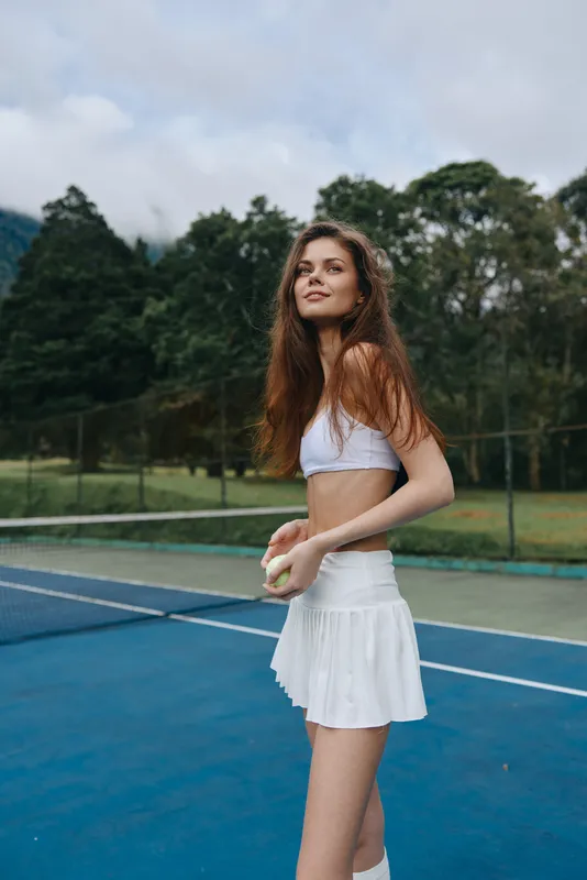 Beautiful athletic woman playing tennis in a white outfit on an outdoor court surrounded by greenery, exuding confidence and grace while holding a tennis ball