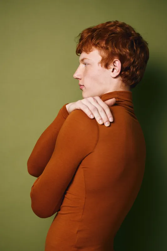 Portrait of young man with red hair wearing brown turtleneck posing with crossed arms against green background. Side view of thoughtful male model in studio light.