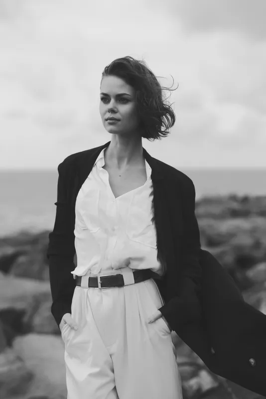 Woman in a blazer stands along a windswept coast in monochrome tones, exuding fashion elegance and bold attitude as the ocean horizon frames a stylish outdoor scene