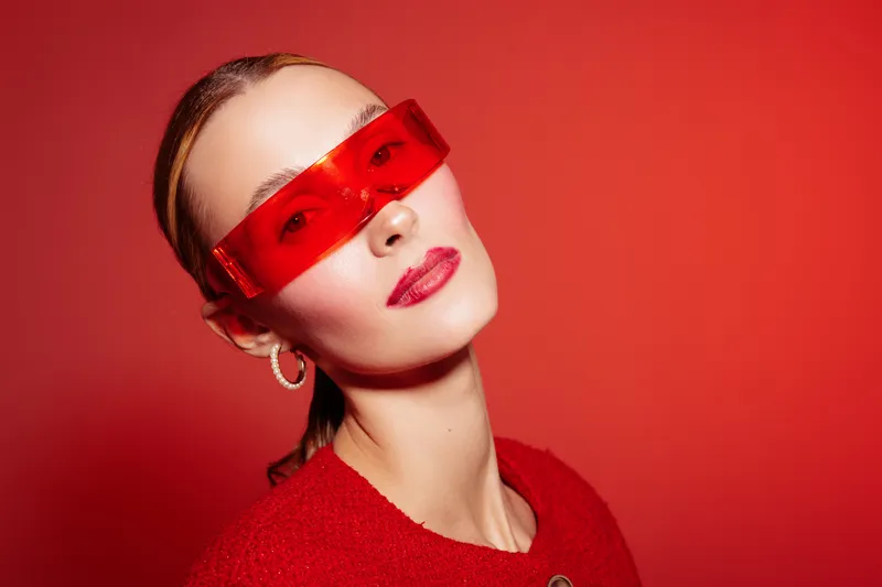 Stylish woman with red sunglasses and red lipstick poses against red background wearing red sweater and hoop earrings. Fashion portrait with vibrant color contrast and modern accessories.
