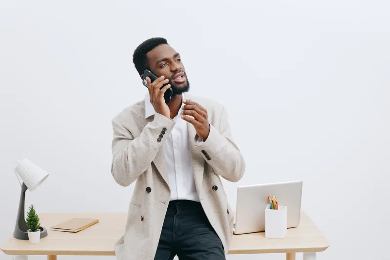 Confident young African American man in beige blazer standing at modern office desk talking on mobile phone, looking serious and professional, with laptop, potted plant and notepad on white background