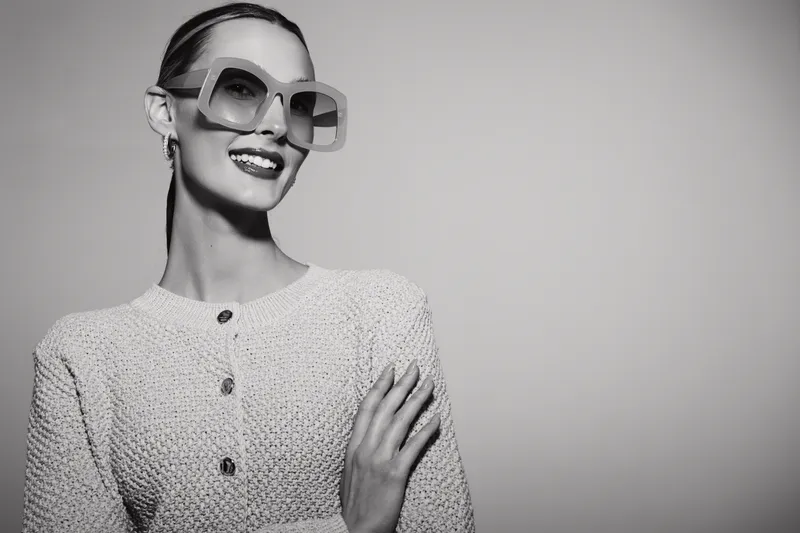 Elegant Caucasian woman with fair skin smiles confidently wearing large sunglasses and textured cardigan. Studio portrait on neutral background. Concept of fashion and style.