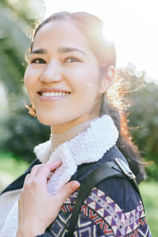 Woman with a warm smile in a sunny outdoor setting, wearing a patterned jacket and fluffy scarf, showcasing casual fashion and natural light in a park