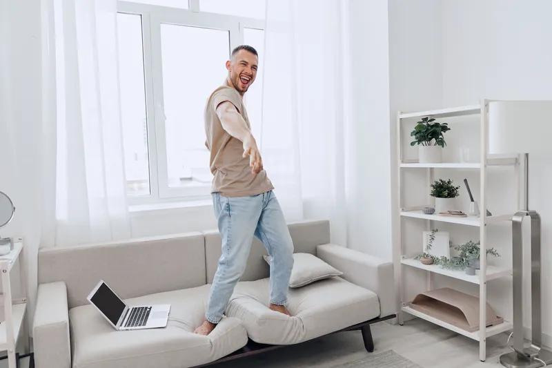 Cheerful man dancing on a couch at home, showcasing a modern, stylish interior with bright, natural light and a minimalist design aesthetic that promotes happiness and comfort.