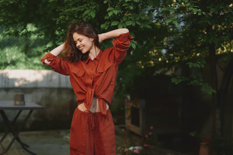 Smiling woman in rust red casual outfit outdoors in a garden. Relaxed pose with hands in her hair. Bright natural light highlights lifestyle, wellness, and fashion vibes