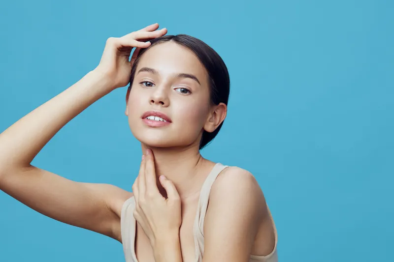 Young model with natural beauty posing against a light blue background, showcasing confidence and grace in a minimalist outfit, emphasizing healthy skin and youthfulness