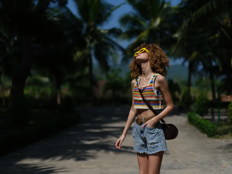 Young woman wearing colorful top and denim shorts enjoys sunny tropical vacation with palm trees and bright yellow sunglasses in summer atmosphere