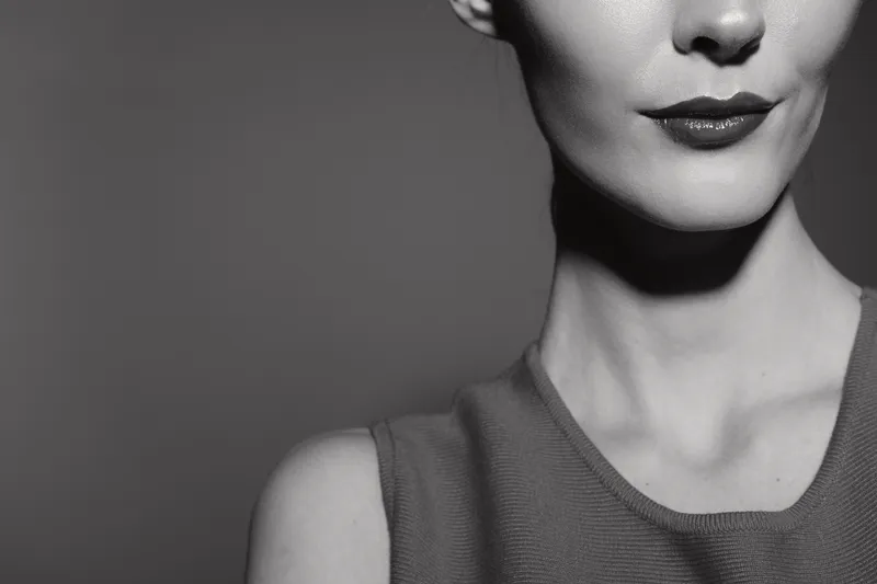 Close up of a white woman with fair skin and red lips wearing a sleeveless top in soft studio lighting. Concept for beauty, portrait, elegance, and minimalism in black and white photography.