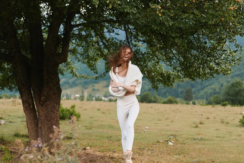 Young woman with long hair stands under a large tree in a field wearing white casual clothes enjoying a windy day with a joyful expression.