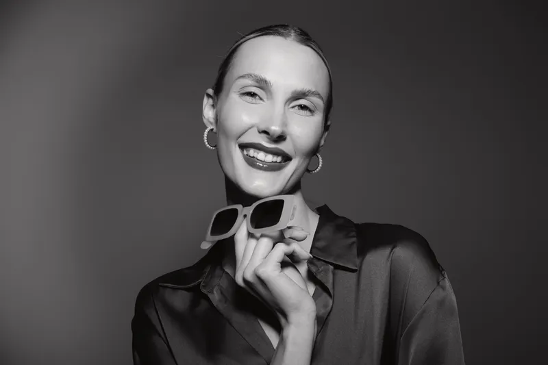 Smiling caucasian woman with pale skin holds sunglasses close to her face, wearing hoop earrings and dark sheer blouse. Concept of fashion and happiness.