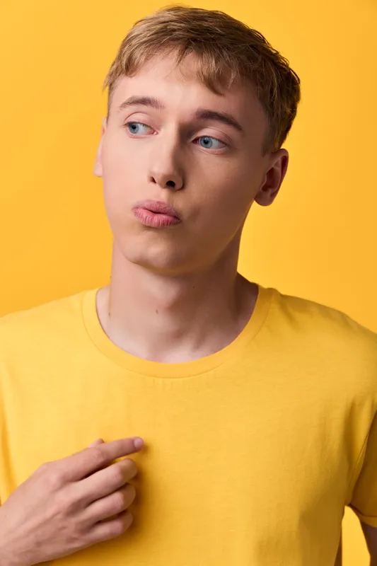Young man in a yellow shirt stands against a bright yellow background, displaying a pensive expression and casual style in a clean studio shot, perfect for lifestyle, fashion, emotion, and concept