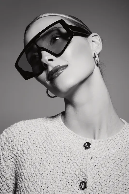 Stylish white woman with light skin wears oversized sunglasses and hoop earrings, dressed in textured sweater. Black and white portrait with elegant fashion concept and confident mood.