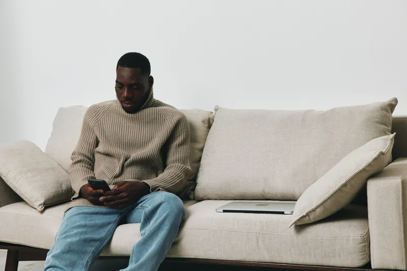 Young Black man sitting on a beige couch, checking his phone while relaxed, portraying a sense of calm and modern lifestyle in a minimalist environment