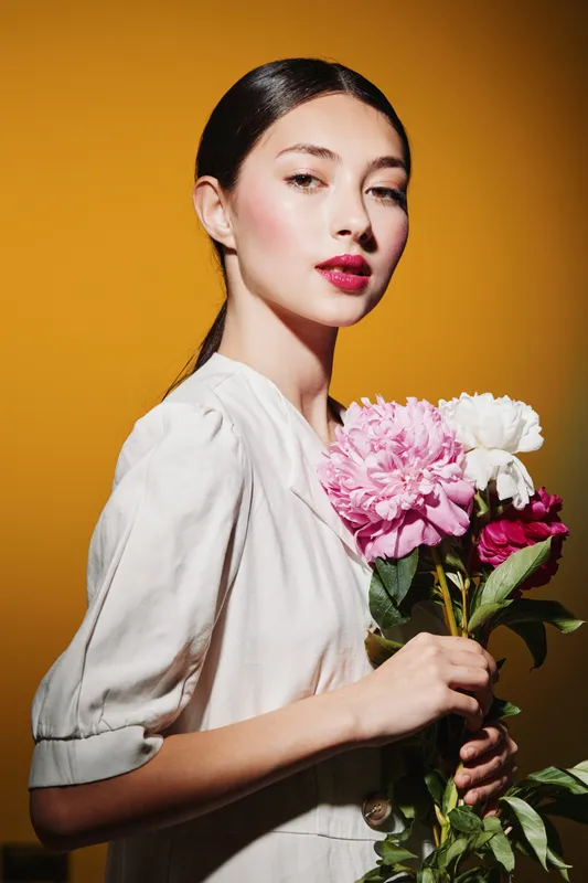 Elegant young woman with polished makeup and vibrant lipstick holding fresh pink and white flowers against a warm amber background in a fashion beauty creative shoot.