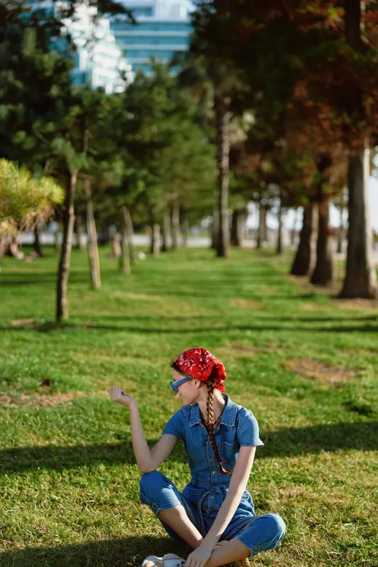 lifestyle woman in modern boho-western denim street style sitting on grass, wearing red bandana and sunglasses, film color effect emphasizes relaxed outdoor mood