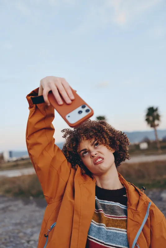 Woman outdoors lifestyle selfie smartphone curly hair jacket, smiling in warm daylight, holding phone near face, relaxed posture, orange tones, palm trees and hills blurred background