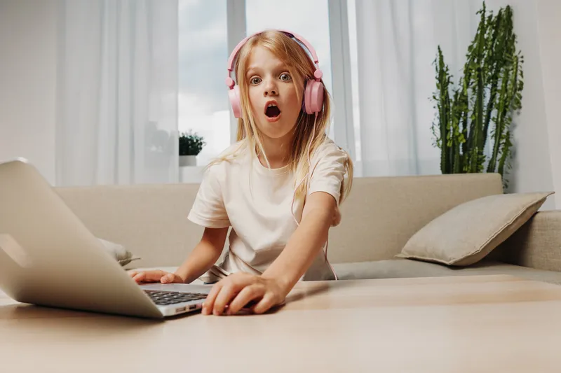 Surprised girl with headphones uses laptop at home, sitting on couch in bright room. Concept of online learning, digital communication, and children's curiosity.