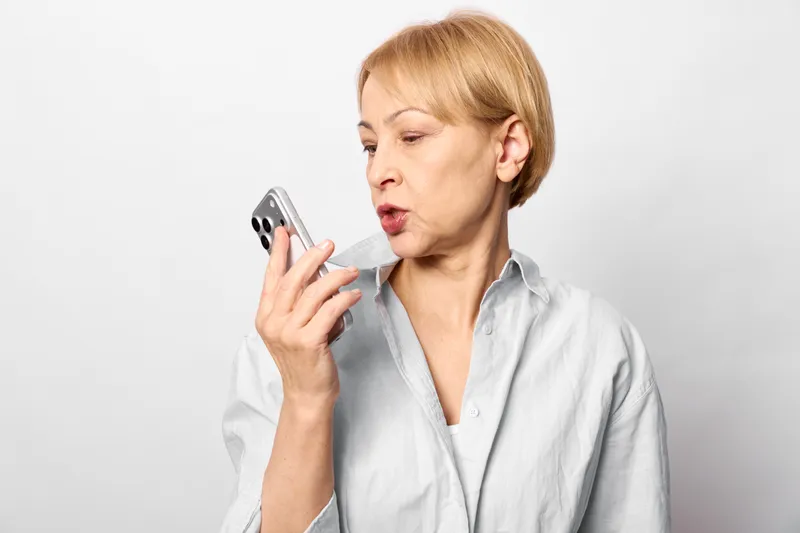 Anxious older woman holds smartphone to ear, mouth open in shock, surprised, isolated on white background, concept of anxiety and stress, phone call crisis