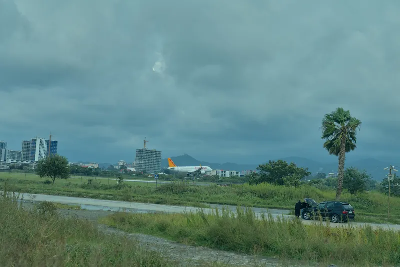 Airfield landscape with distant aircraft, roadside car, open field and cloudy sky, capturing travel and aviation atmosphere. A moody overcast scene shows greens, a parking area, and a distant town on