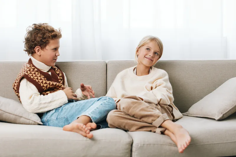 Two children relax on a soft sofa under natural light, sharing a playful moment in cozy sweaters as they chat and enjoy quiet companionship.